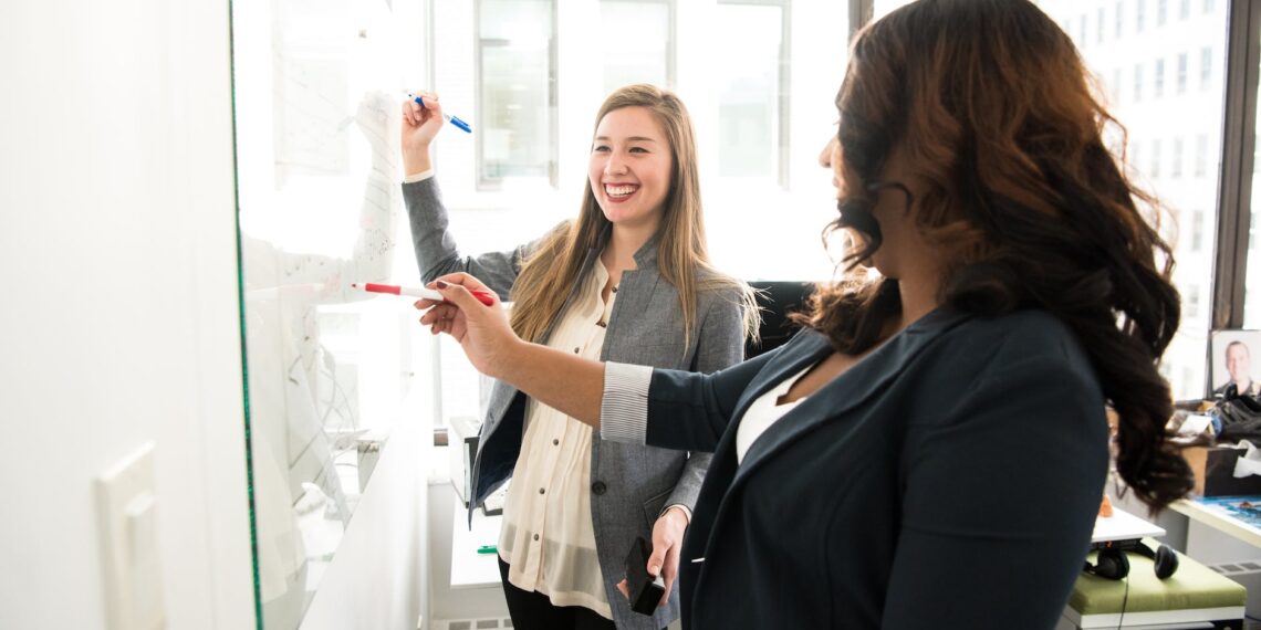 Two women engaged in workplace conflict standing in front of a whiteboard in an office.