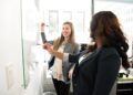 Two women engaged in workplace conflict standing in front of a whiteboard in an office.