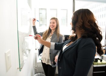 Two women engaged in workplace conflict standing in front of a whiteboard in an office.