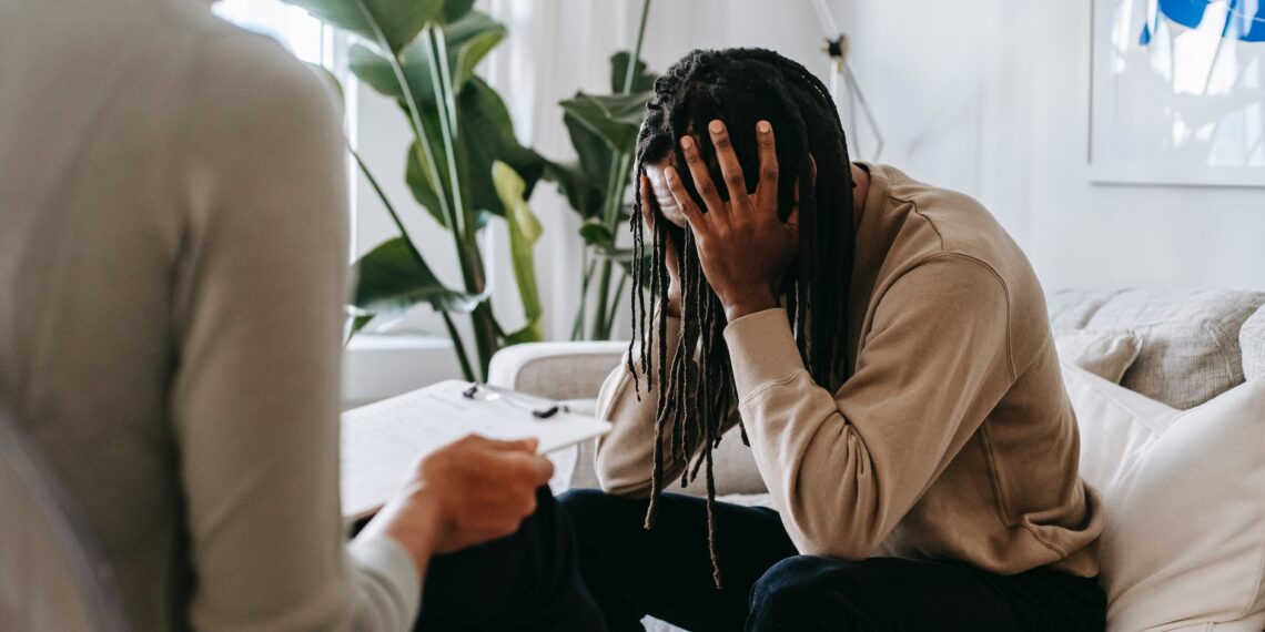 A woman is sitting on a couch, visibly distressed with her head in her hands, displaying signs of attachment trauma.