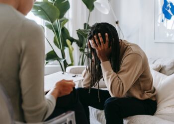 A woman is sitting on a couch, visibly distressed with her head in her hands, displaying signs of attachment trauma.