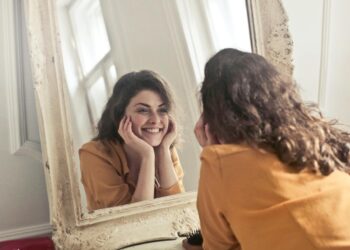 A woman with curly hair and a yellow shirt smiles at her reflection in a large ornate mirror, taking a moment to acknowledge her feelings and understand why she worries about the future.