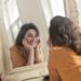 A woman with curly hair and a yellow shirt smiles at her reflection in a large ornate mirror, taking a moment to acknowledge her feelings and understand why she worries about the future.