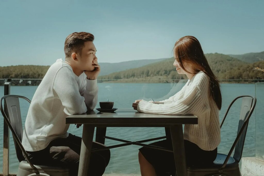 Two people sit across from each other at a table by a lake, engaged in conversation. They are both wearing white tops, and the scenic background features mountains and a clear sky.