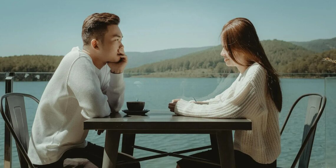 Two people sit across from each other at a table by a lake, engaged in conversation. They are both wearing white tops, and the scenic background features mountains and a clear sky.