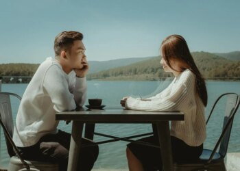 Two people sit across from each other at a table by a lake, engaged in conversation. They are both wearing white tops, and the scenic background features mountains and a clear sky.