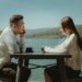 Two people sit across from each other at a table by a lake, engaged in conversation. They are both wearing white tops, and the scenic background features mountains and a clear sky.