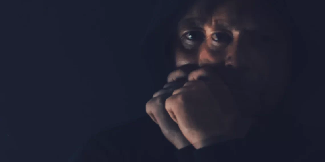 A person in a hooded sweatshirt holds their hands to their face in a dimly lit setting, embracing the power of silence.