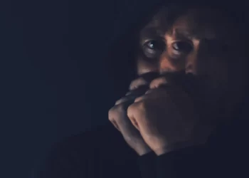 A person in a hooded sweatshirt holds their hands to their face in a dimly lit setting, embracing the power of silence.